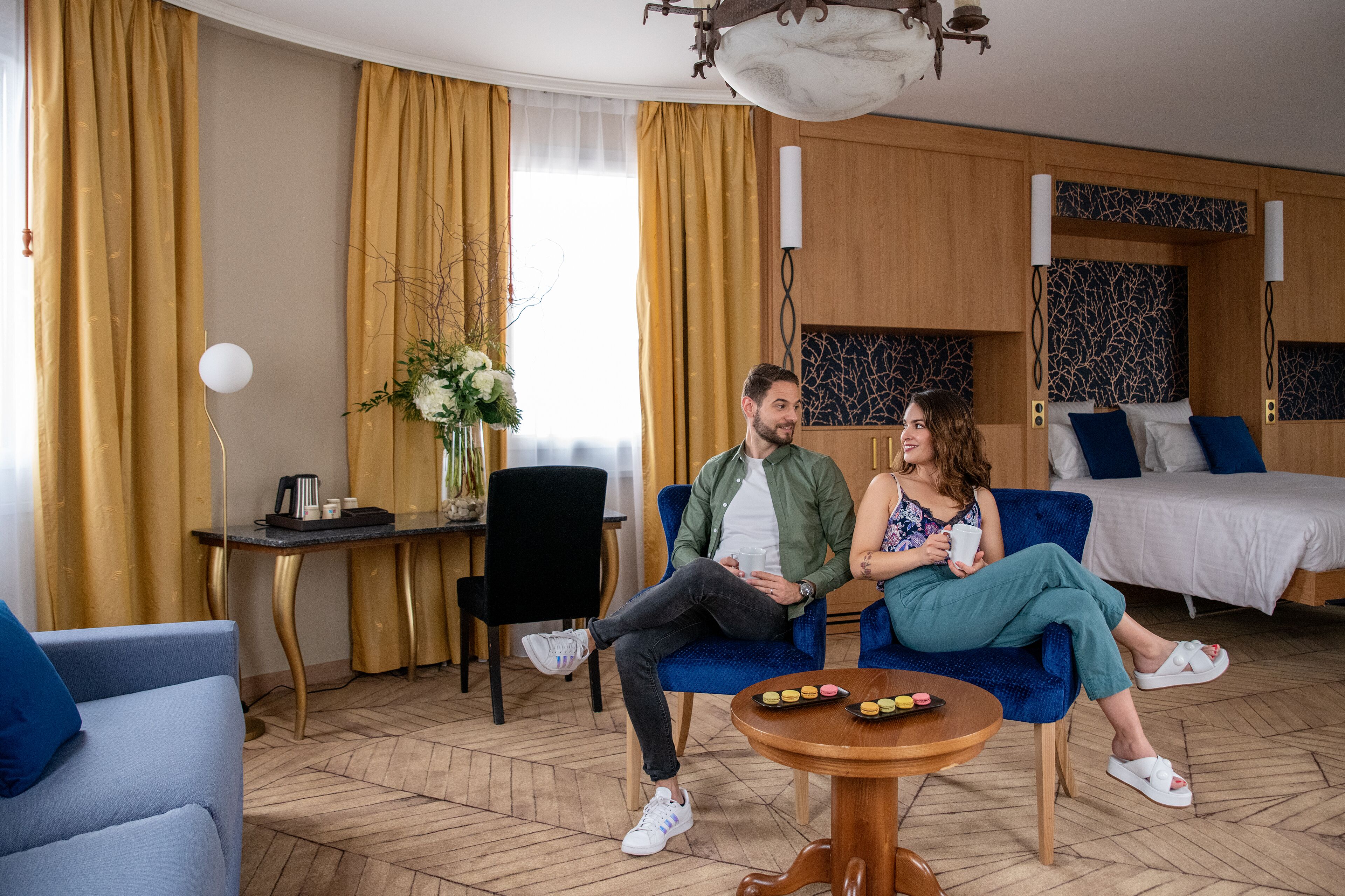 A hotel room with a bed, a blue sofa, a round wooden table with snacks, a small side table, and a vase with flowers.