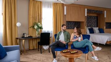 Una habitación de hotel con una cama, un sofá azul, una mesa redonda de madera con aperitivos, una mesita auxiliar y un jarrón con flores.