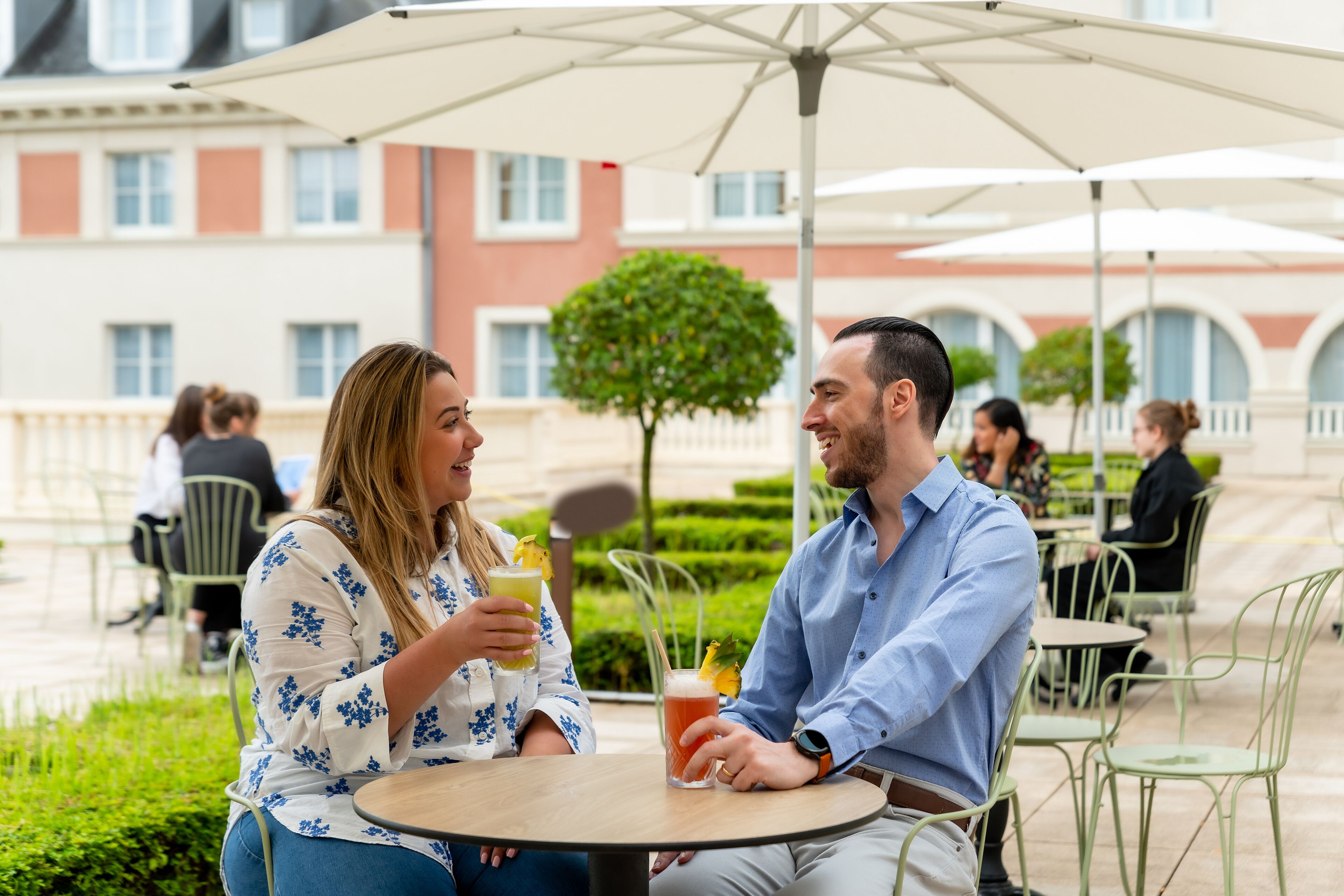 Two people sitting at an outdoor table with drinks, surrounded by green chairs and umbrellas, with a building and a tree in the background.