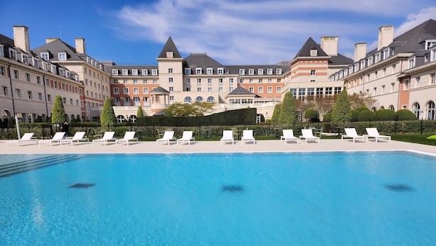 A swimming pool with white lounge chairs, surrounded by a building with multiple balconies and a well-maintained hedge.