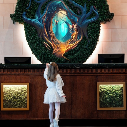 A young girl in a white dress standing in front of a wall with a large, illuminated tree mural.