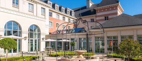 Un patio con mobiliario exterior, una fuente y un edificio de varios pisos con ventanas grandes.