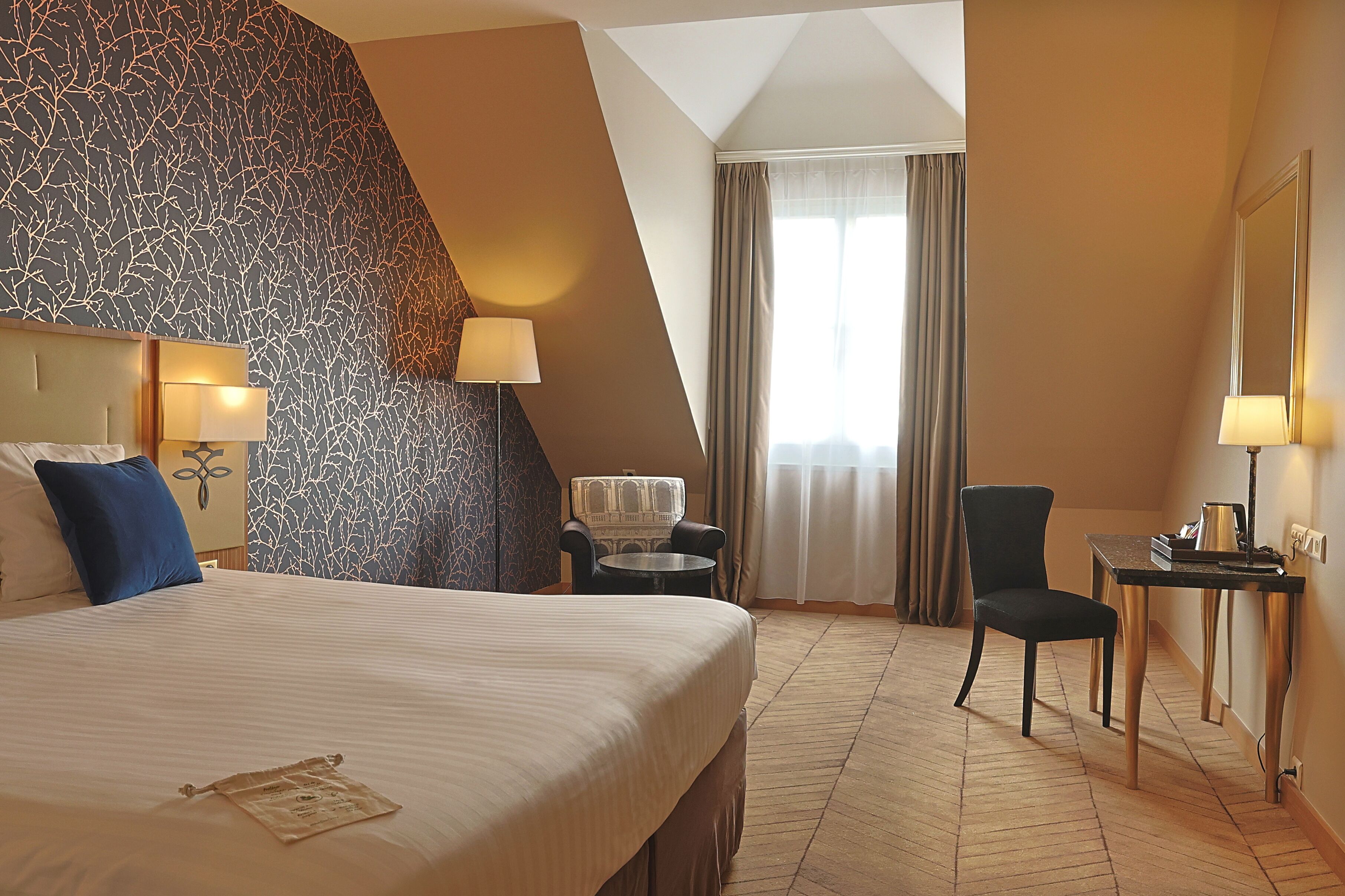A hotel room with a large bed, a chair, a small table, and a desk with a lamp.