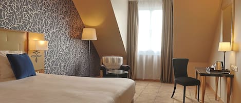 A hotel room with a large bed, a chair, a small table, and a desk with a lamp.