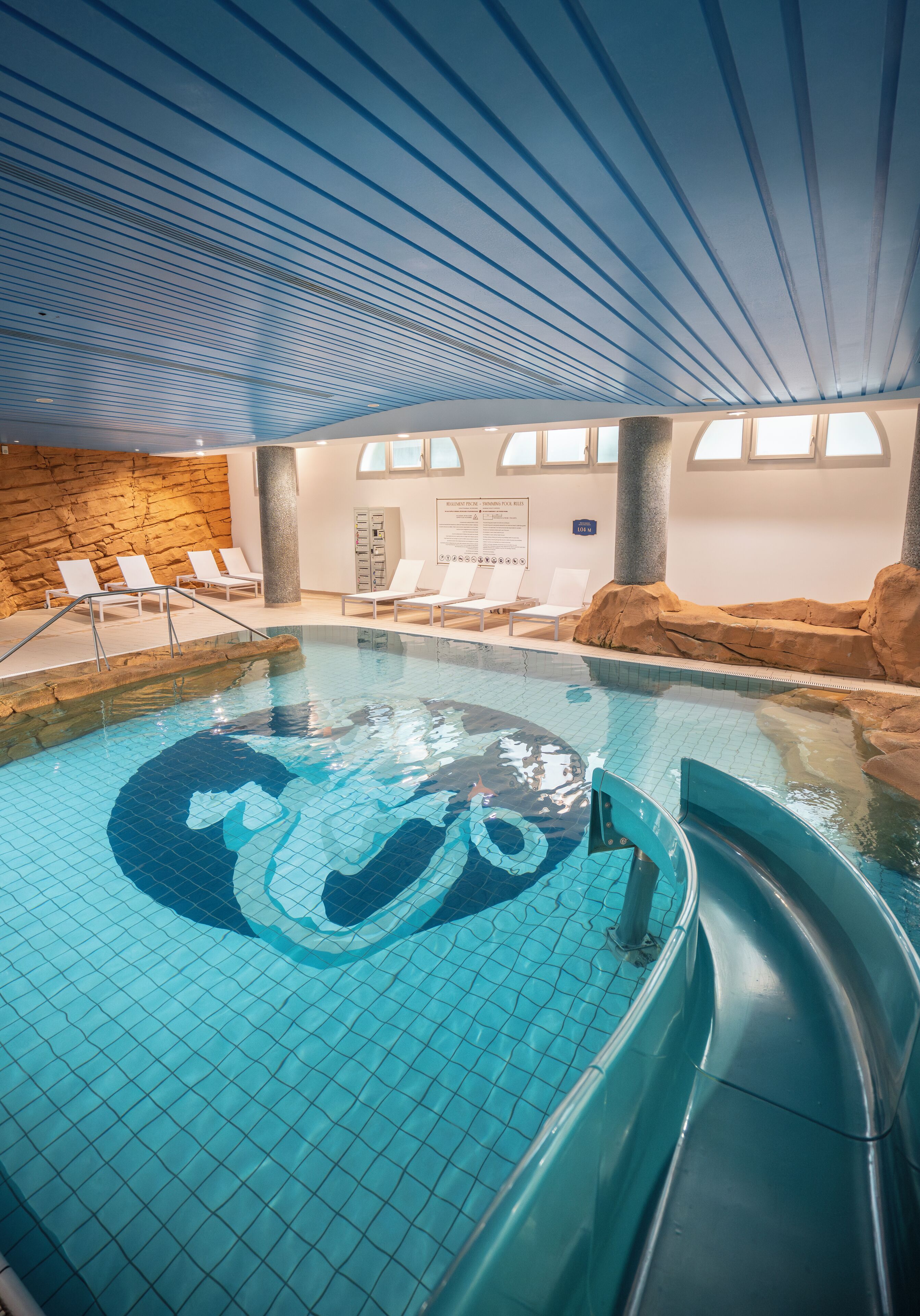 An indoor swimming pool with a clear water feature, lounge chairs, and a stone wall.