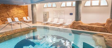 An indoor swimming pool with a clear water feature, lounge chairs, and a stone wall.