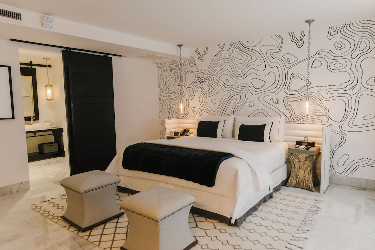 A modern bedroom with a large bed, two ottomans, a nightstand, and a wall with a unique black and white pattern.