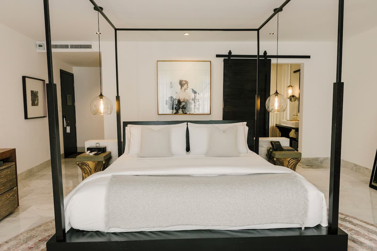A modern hotel room with a large bed, a bedside table, a mirror, and a framed artwork on the wall.