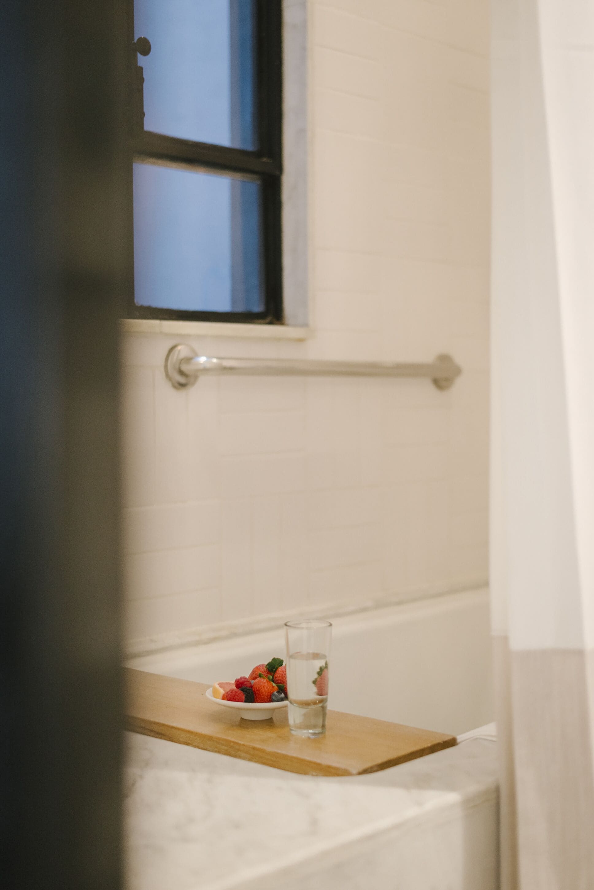 Un baño con una bañera blanca, una ventana y una bandeja de madera que sostiene un cuenco de fruta y un vaso de agua.