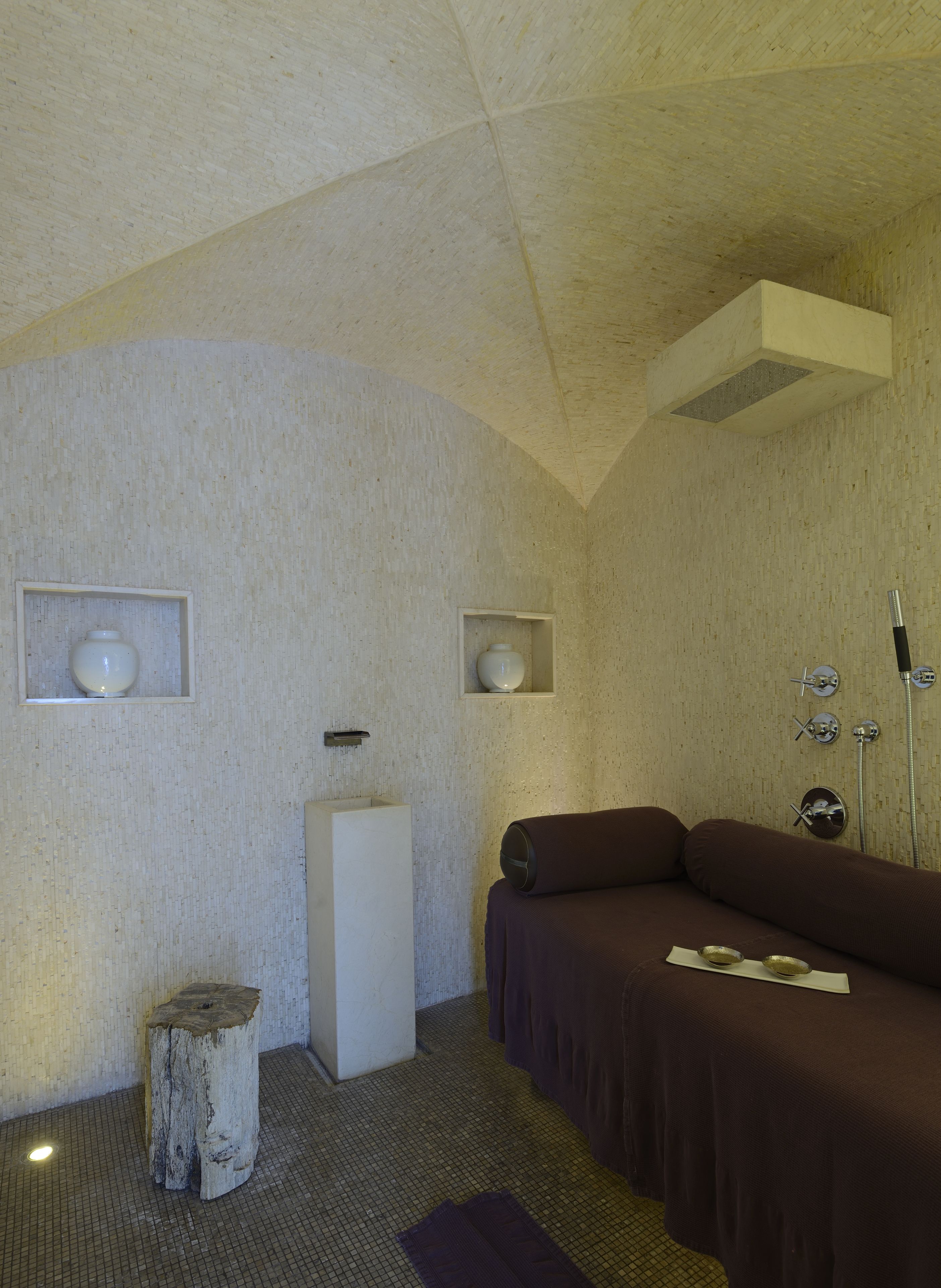 A room with a high ceiling, a large window, a bed, a small table, and a wall-mounted shelf.