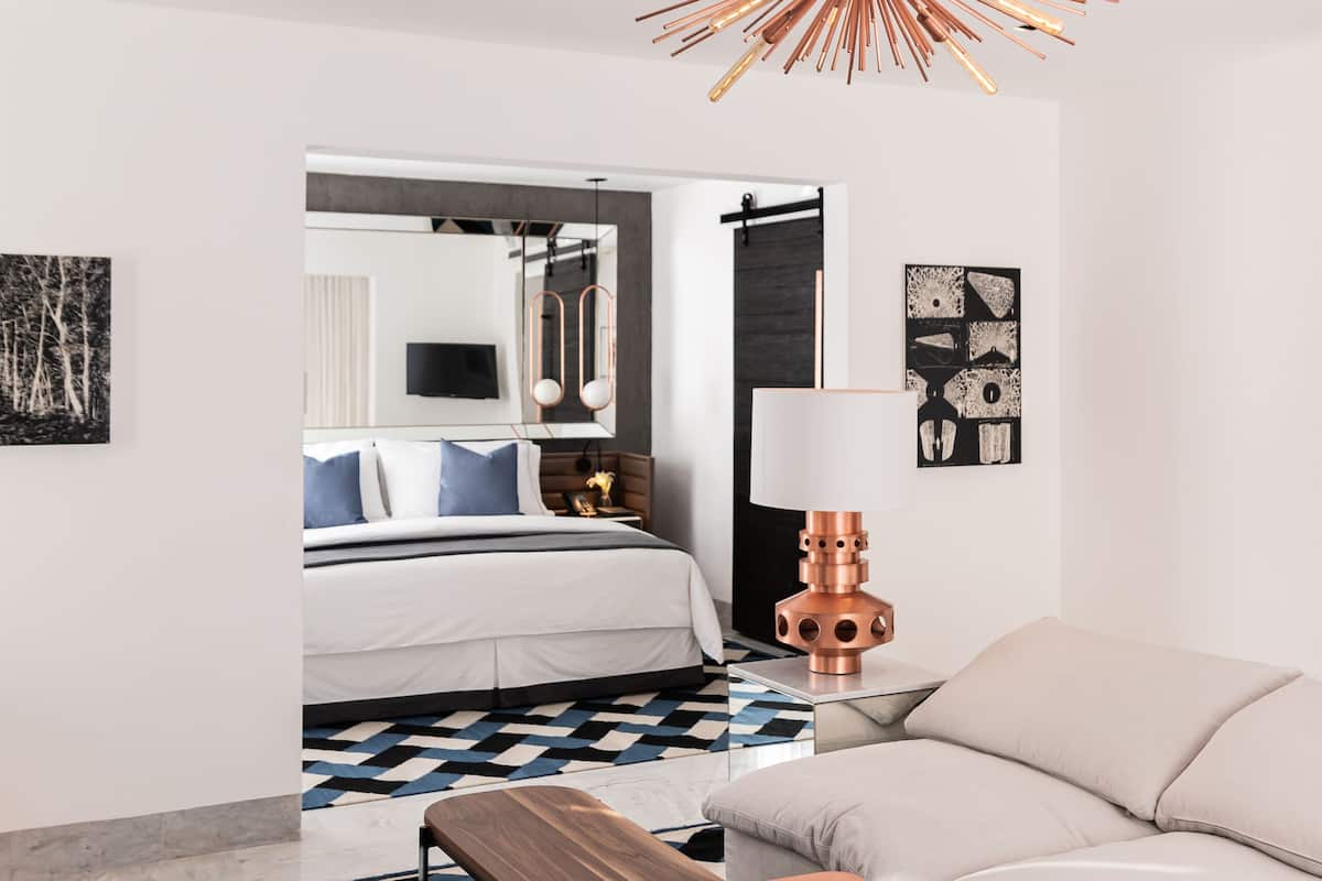 A modern hotel room with a bed, a sofa, a coffee table, a patterned rug, and a unique ceiling light fixture.