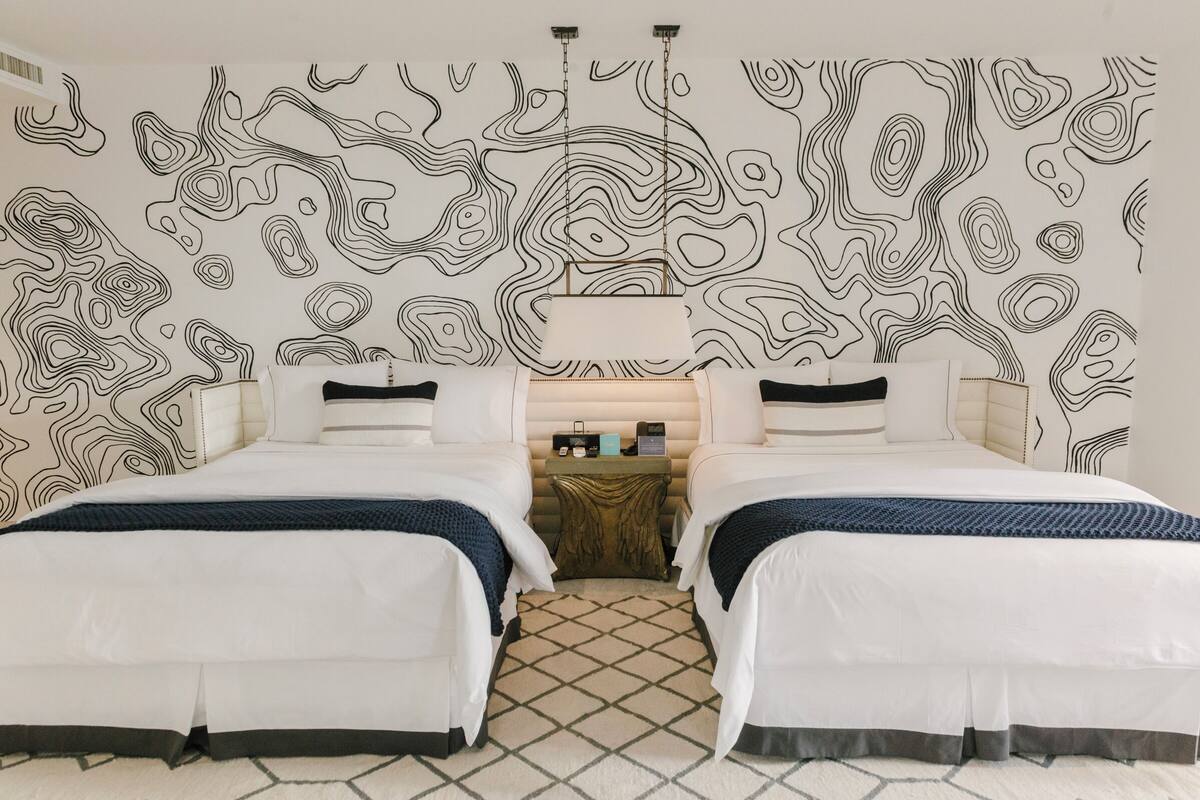 A hotel room with two beds, a patterned wall, a small table with a lamp, and a geometric rug.