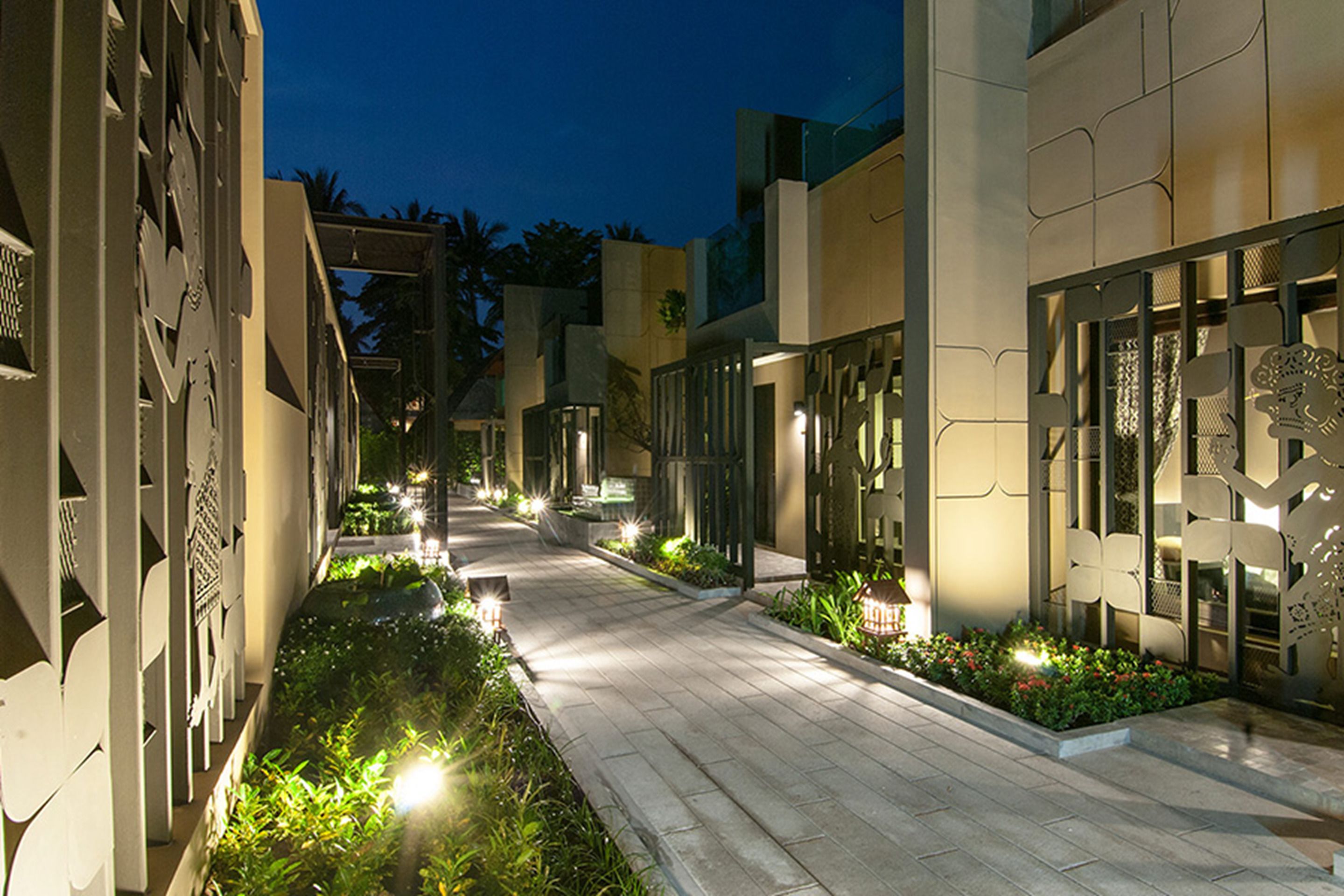 A well-lit pathway flanked by modern buildings with geometric patterns and greenery.
