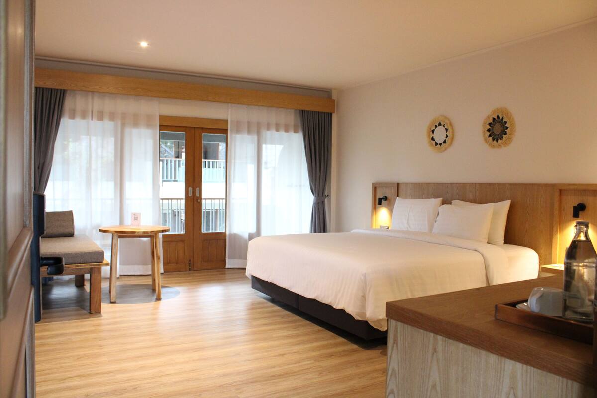 A hotel room with a large bed, a wooden nightstand, a small table, and a sofa. There are two curtains on the window, and a balcony is visible through the window.