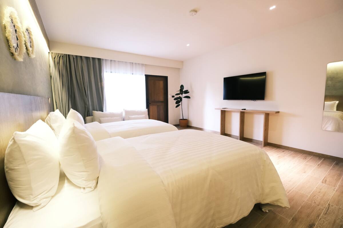 A hotel room with two beds, a flat-screen TV, a potted plant, and a large window with curtains.