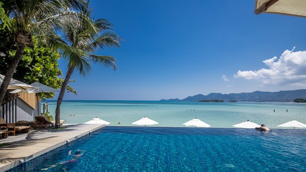 A clear blue pool with a view of the ocean and mountains, surrounded by palm trees and umbrellas. - Baan Haad Ngam Boutique Resort & Villas (Koh Samui)