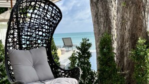 A wicker chair with a cushion, positioned near a tree and overlooking a beach with lounge chairs. - Baan Haad Ngam Boutique Resort & Villas (Koh Samui)
