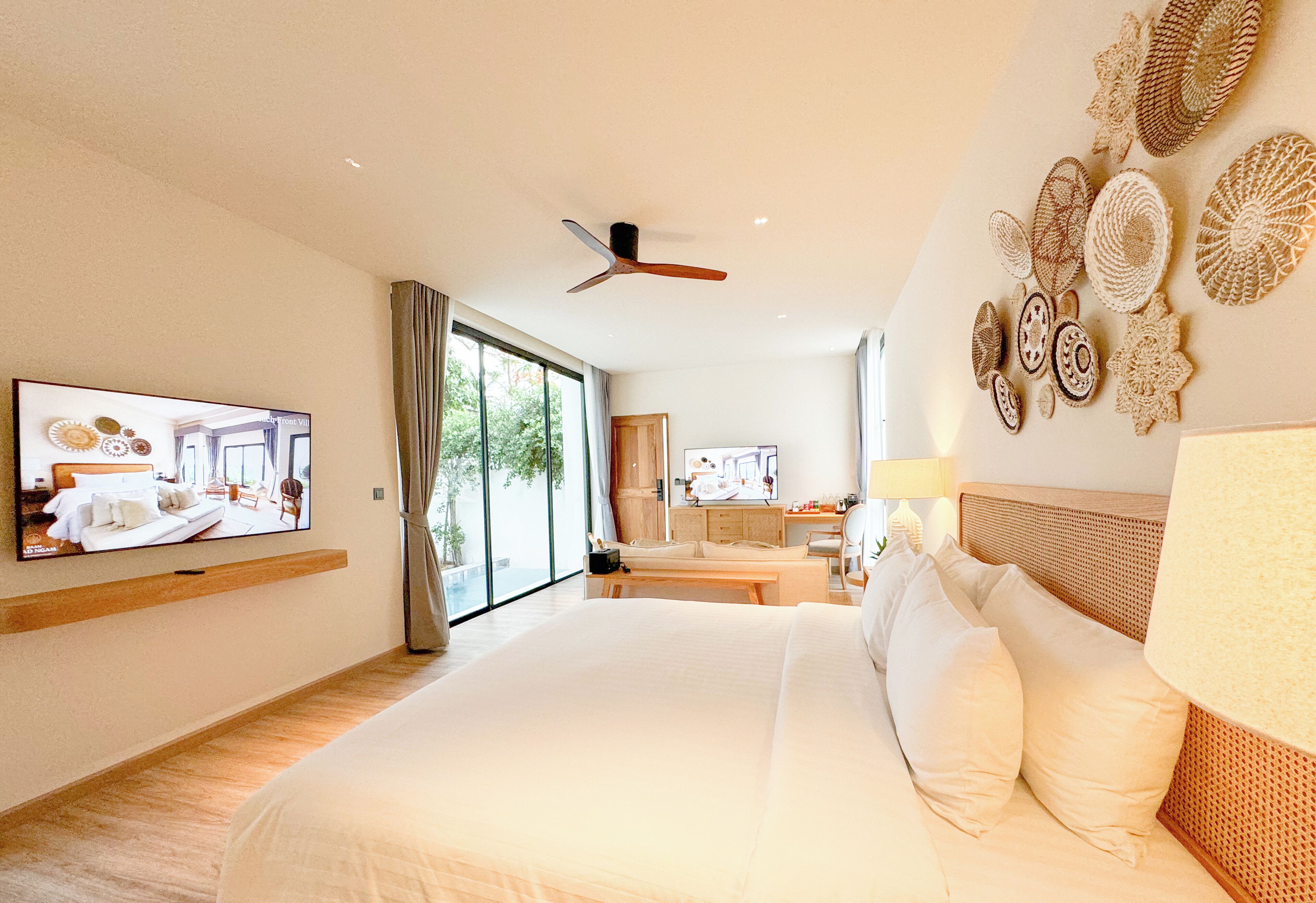 A modern hotel room with a large bed, a television mounted on the wall, a ceiling fan, and a view of the outdoors through the sliding glass door.