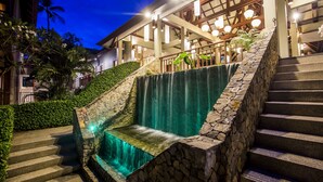 A stone staircase leading to a waterfall, surrounded by lush greenery and a building with large windows and hanging lights. - Baan Haad Ngam Boutique Resort & Villas (Koh Samui)