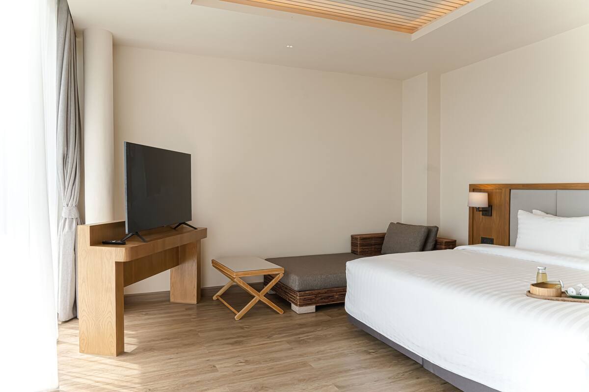 A modern hotel room with a large bed, a desk with a flat-screen TV, a small seating area, and a bedside table with a lamp.