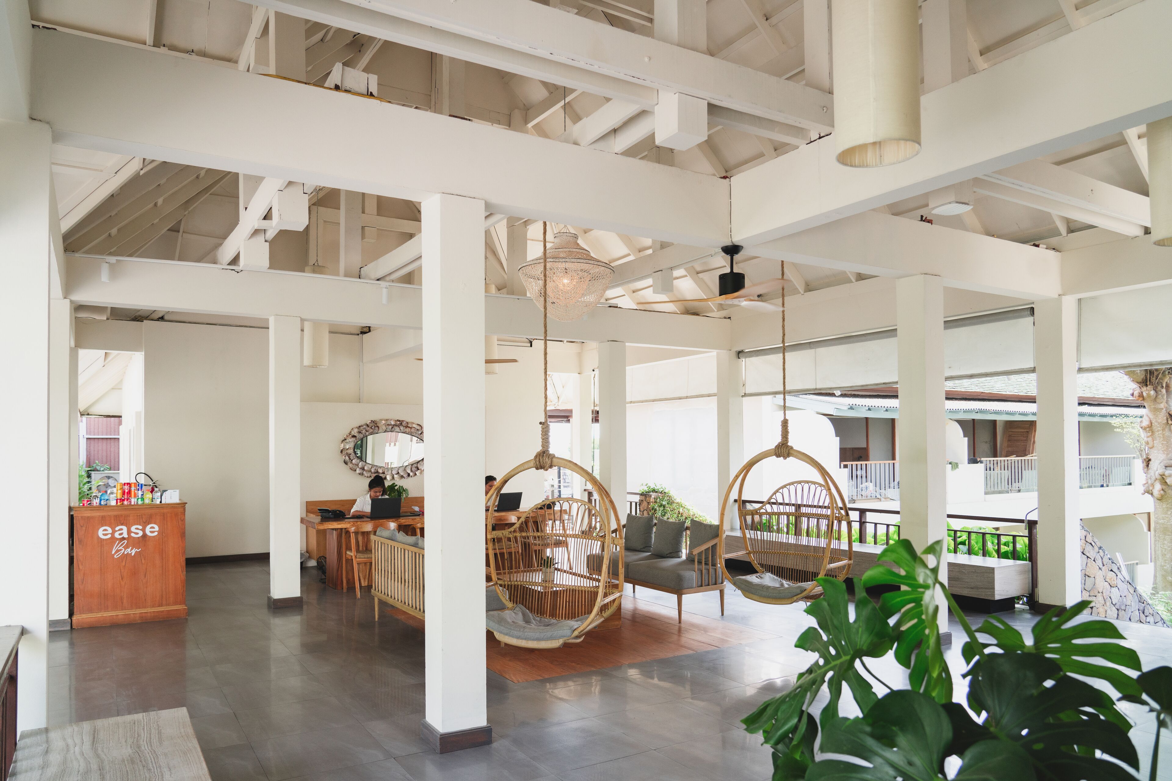 A spacious lobby with high ceilings, exposed beams, and hanging wicker chairs. A reception desk labeled "ease" is visible.