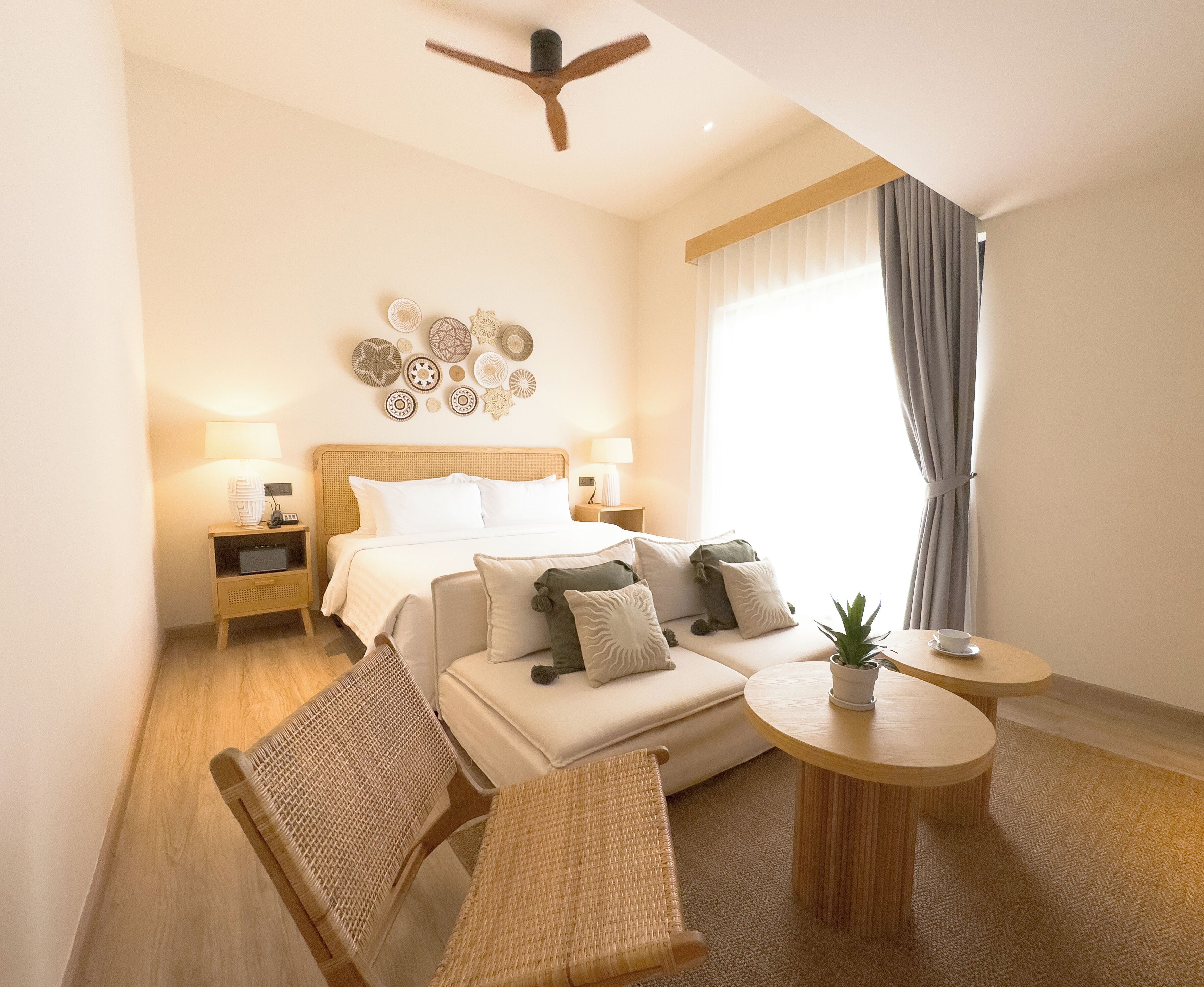 A hotel room with a bed, bedside tables, a sofa, a chair, a small table with a plant, and a ceiling fan.