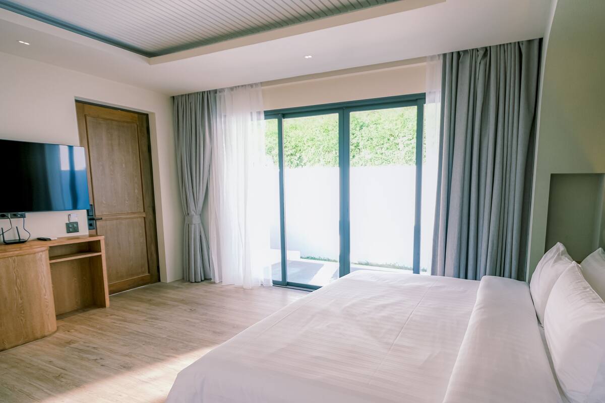 A hotel room with a large bed, a TV on a wooden stand, and a view of greenery through the window.
