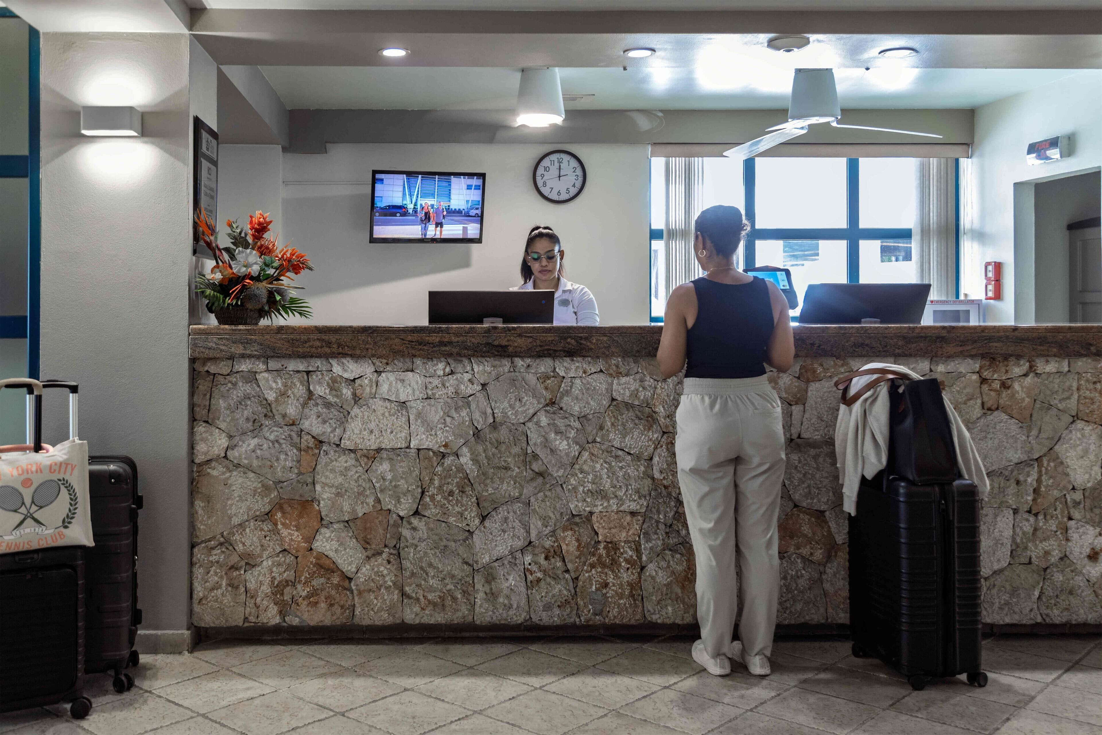 Un mostrador de recepción con pared de piedra, un reloj y una pantalla de televisión mostrando gente.