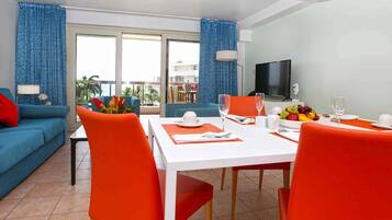 Une chambre d’hôtel moderne dotée d’un coin repas comprenant une table blanche, des chaises orange et un téléviseur fixé au mur.