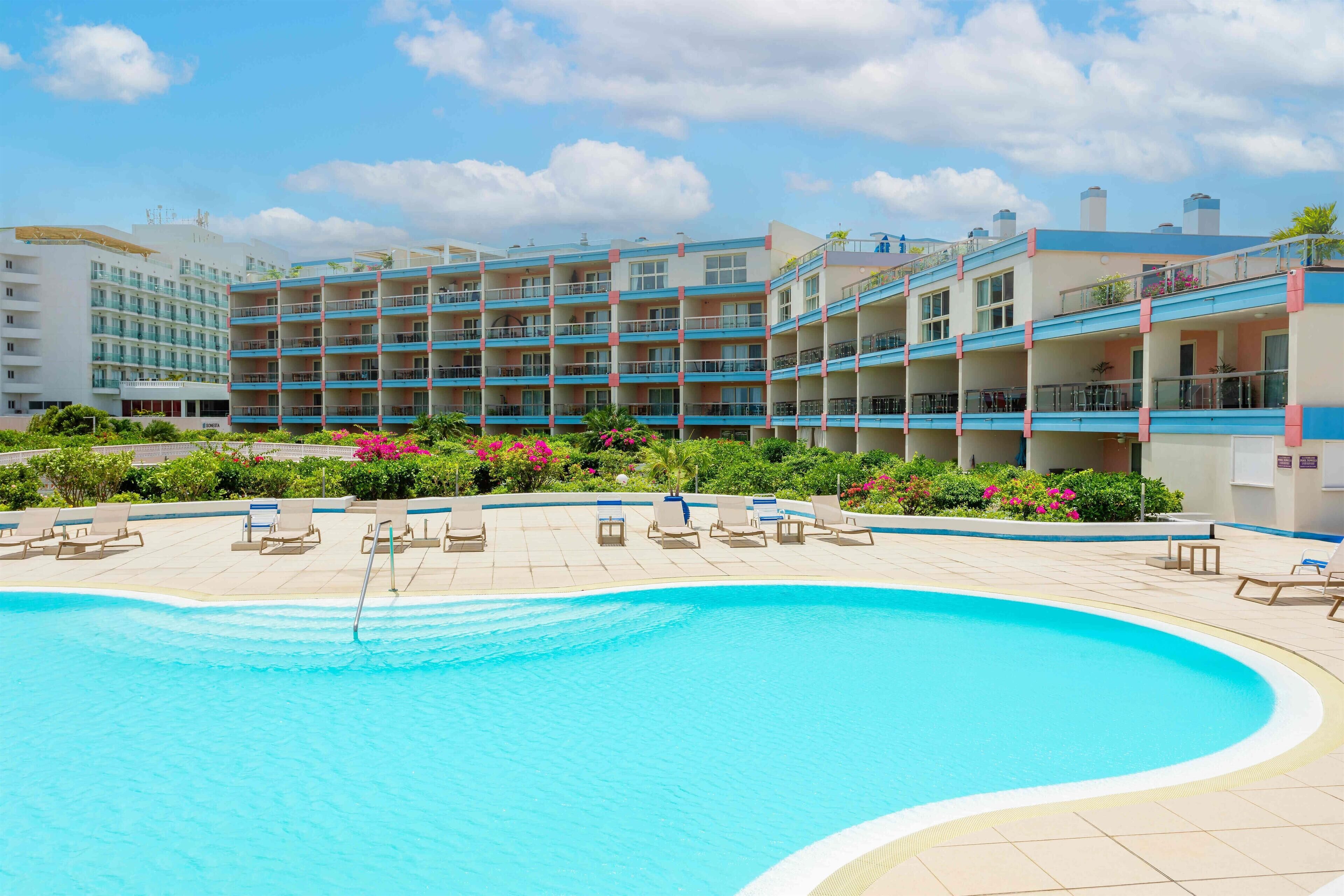 Un hotel con piscina, varios balcones y un jardín bien cuidado con flores rosadas.
