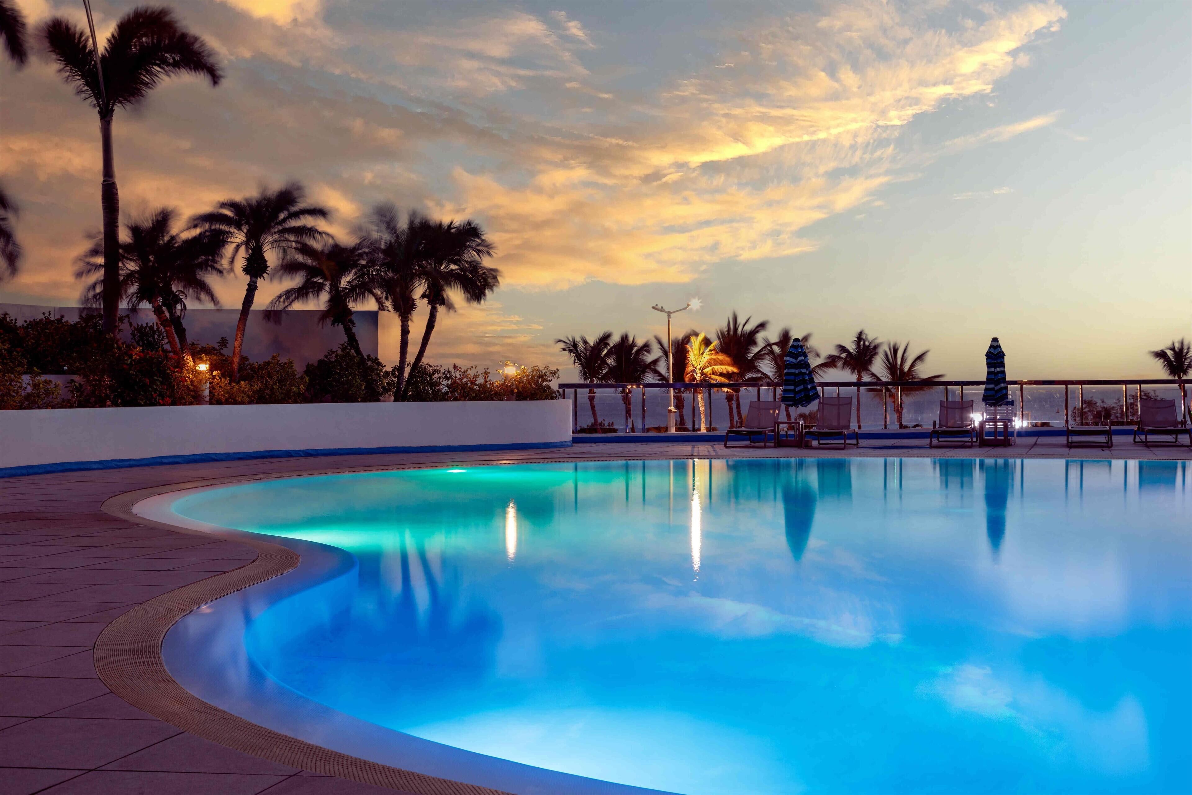 A swimming pool with a curved edge, surrounded by palm trees and lounge chairs, under a sunset sky.