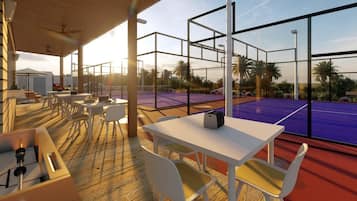 Un área de estar al aire libre con mesas y sillas, una cancha de tenis visible a través de grandes ventanales y una terraza de madera.