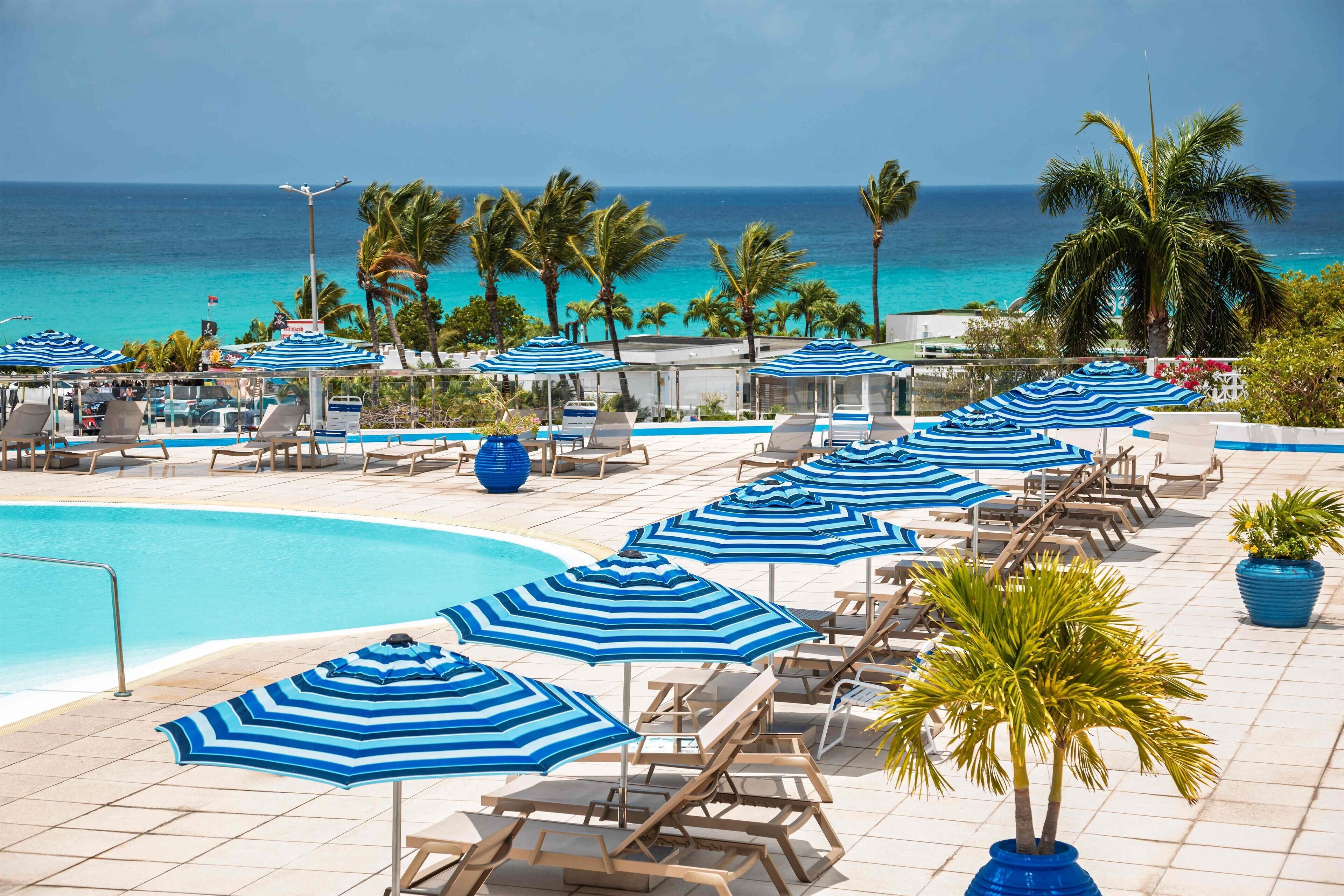 A poolside area with lounge chairs and blue-striped umbrellas, overlooking a clear blue ocean.