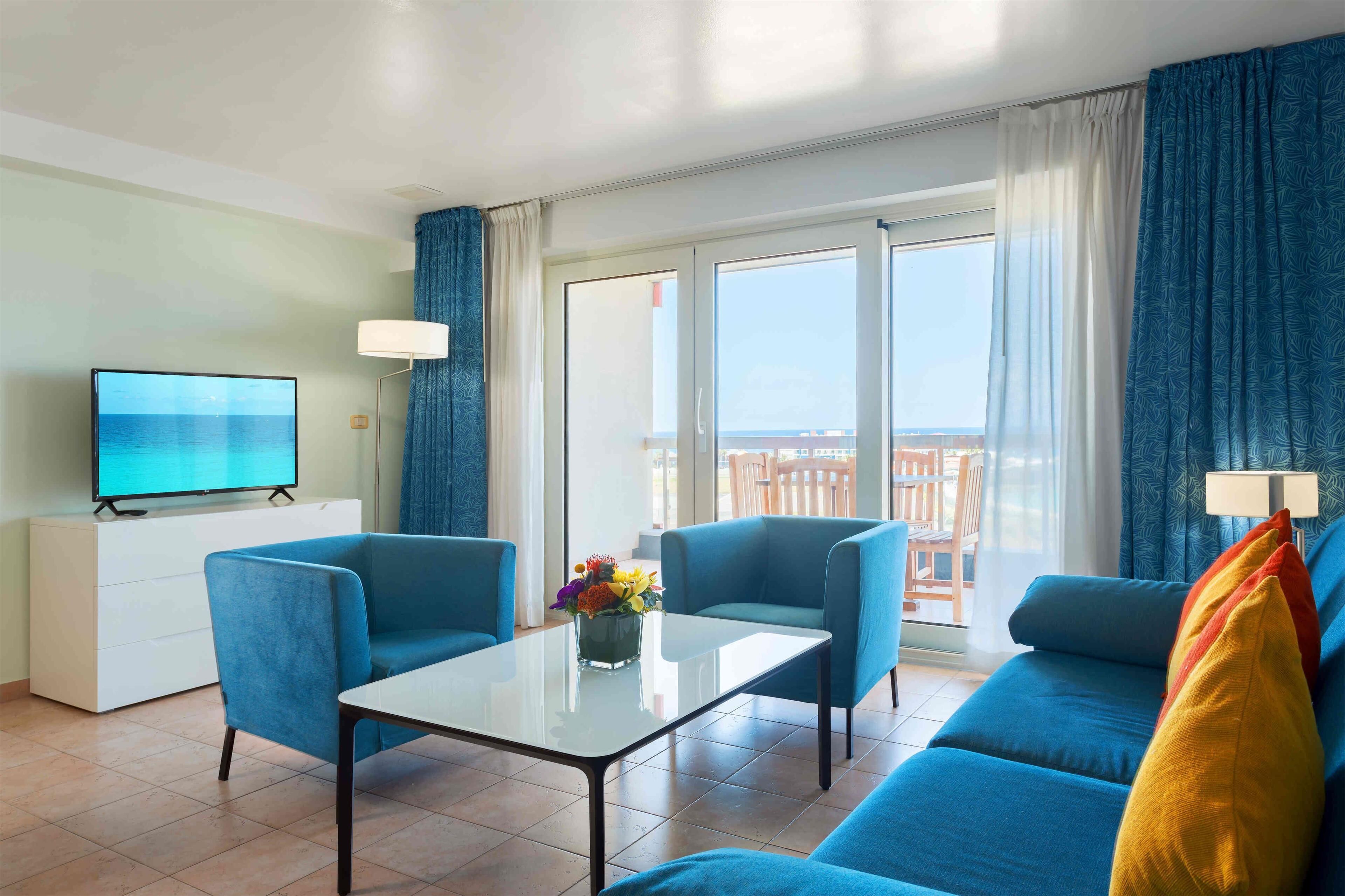 Una sala moderna con un televisor de pantalla plana grande, dos sillones azules, un sofá, una mesa de centro de vidrio y un balcón con vistas.