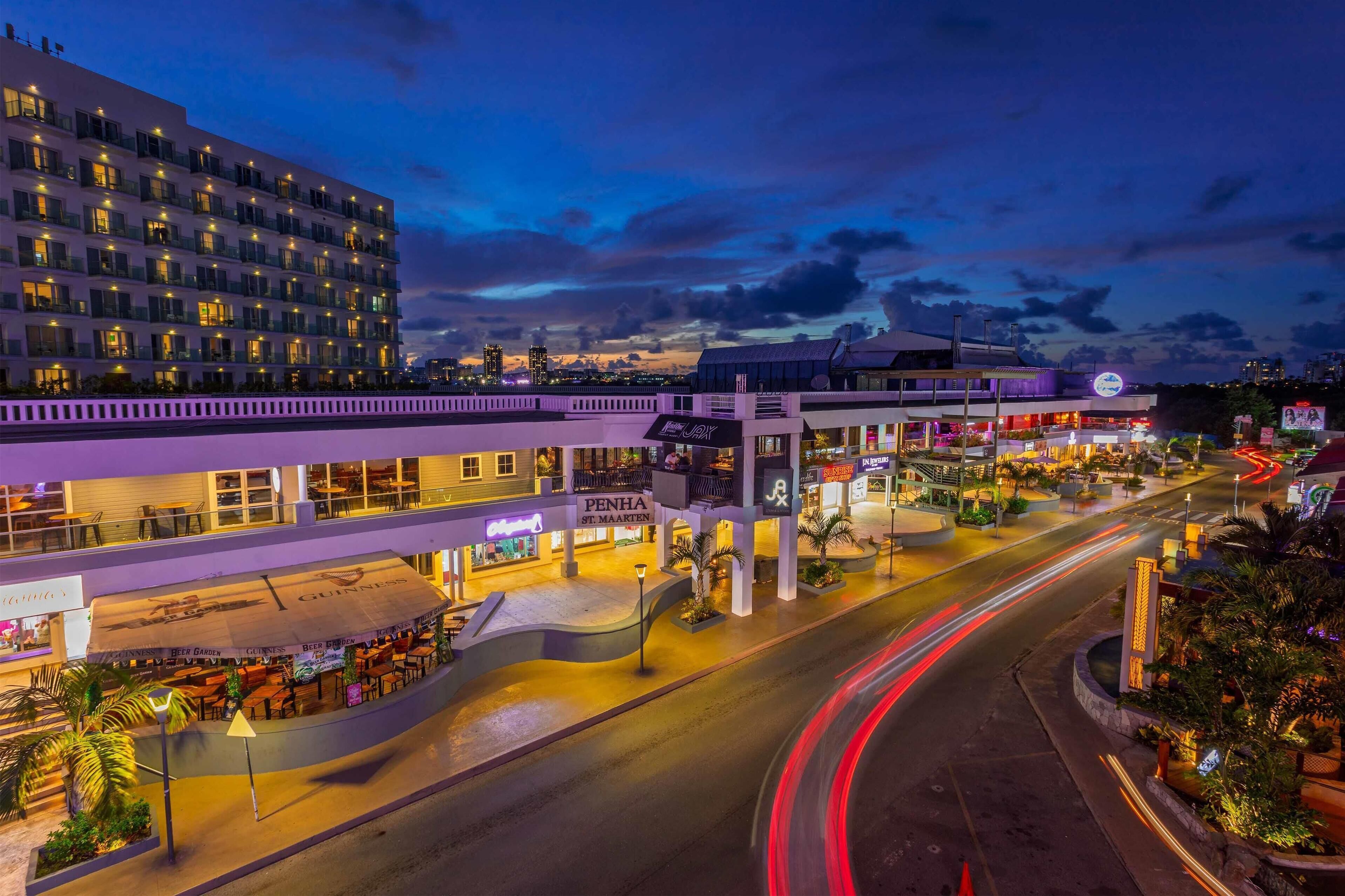 Un hôtel à plusieurs étages au design moderne, avec une enseigne bien visible indiquant « Penha St. Maarten », des espaces de détente en plein air et une rue bien éclairée, agrémentée de jeux de lumière.