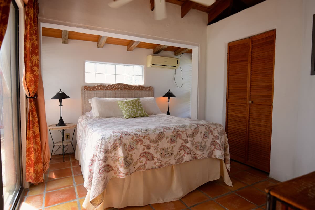 Une chambre à coucher avec un lit, deux lampes de chevet, une fenêtre, un appareil de climatisation et une porte en bois.