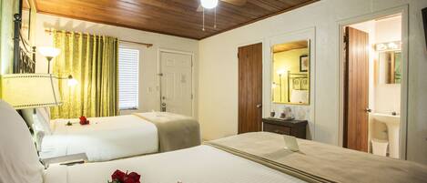 A hotel room with two beds, a ceiling fan, a window with curtains, a bedside table with a lamp, and a bathroom visible through an open door.