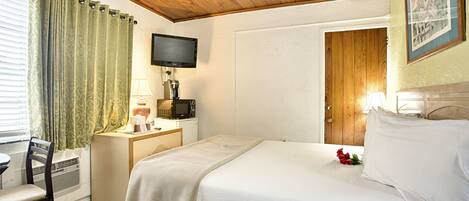 Habitación de hotel con techo de madera, una cama con ropa blanca, un escritorio con lámpara, un televisor y una ventana con cortinas.