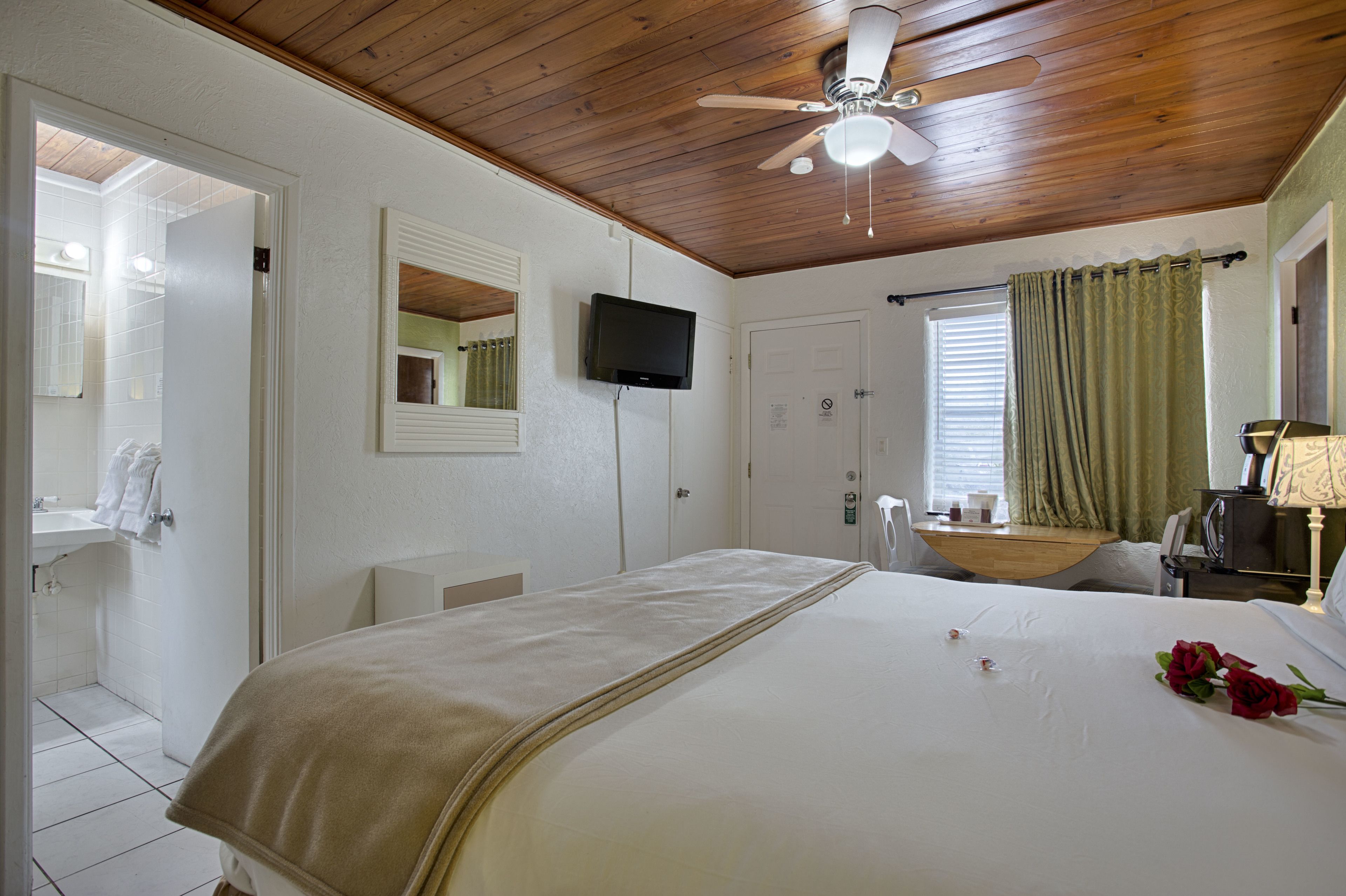 Une chambre d’hôtel équipée d’un lit, d’une télévision, d’un ventilateur de plafond, d’une fenêtre avec des rideaux et d’une salle de bain visible à travers une porte ouverte.