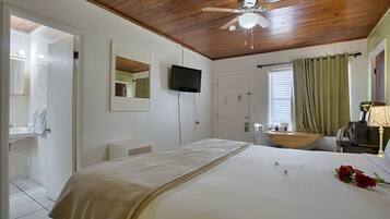 Une chambre d’hôtel équipée d’un lit, d’une télévision, d’un ventilateur de plafond, d’une fenêtre avec des rideaux et d’une salle de bain visible à travers une porte ouverte.