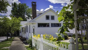 A white house with a brick chimney, a white picket fence, and a garden with yellow flowers. - Homestead Inn (New Milford)