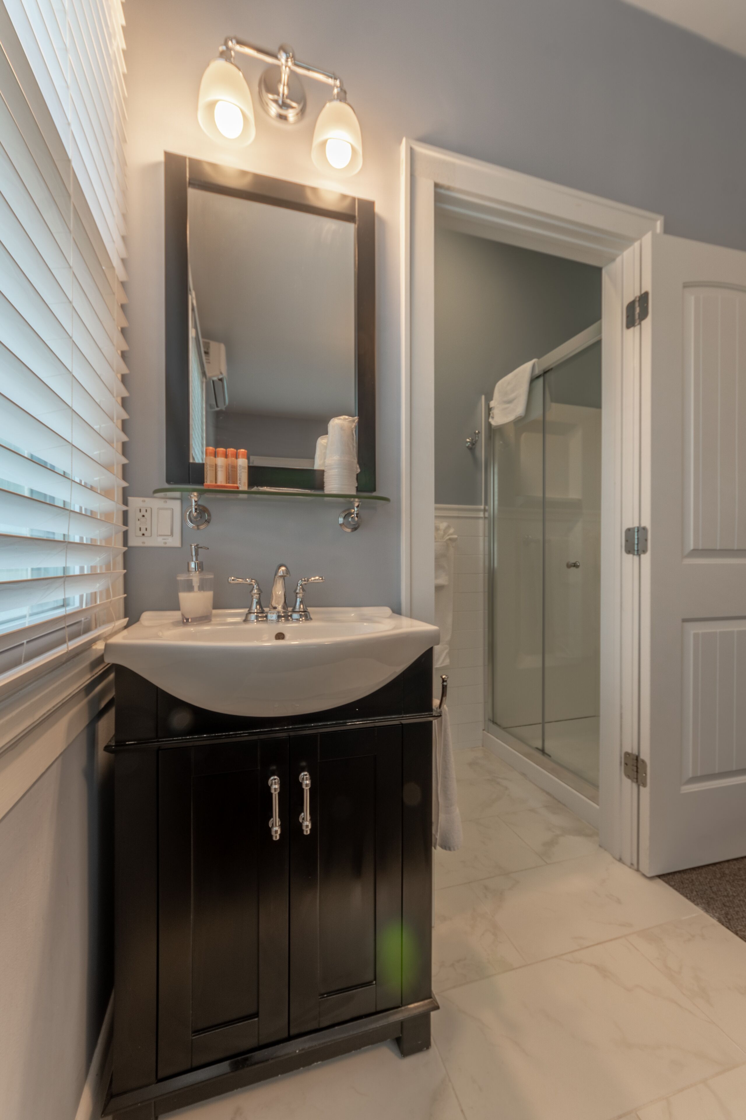 A bathroom with a white sink, dark cabinets, a mirror with lights, and a glass shower door.