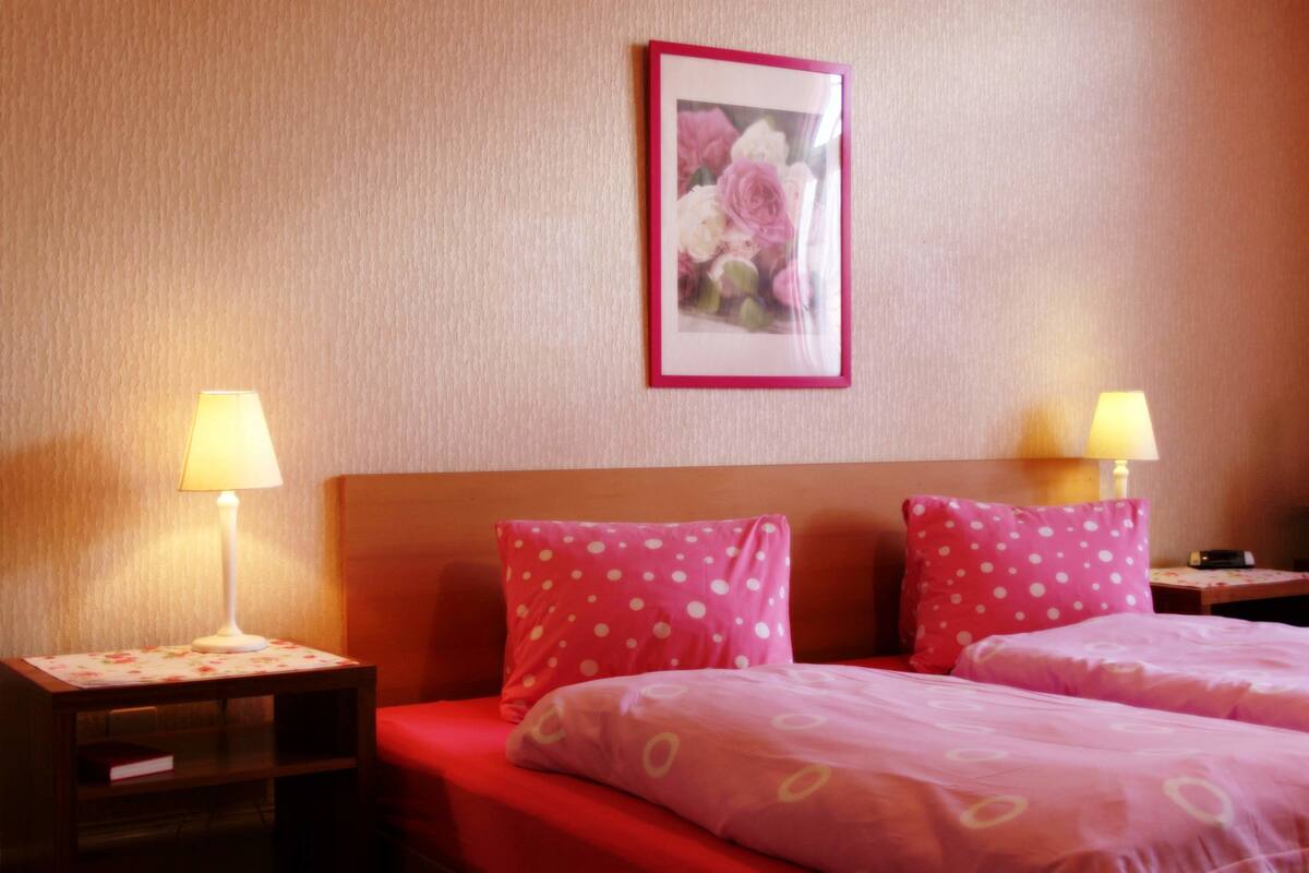 Une chambre d’hôtel avec deux lits, chacun agrémenté de draps roses et d’oreillers blancs à pois. Il y a deux tables de chevet avec des lampes, et un tableau représentant des fleurs est accroché au mur.