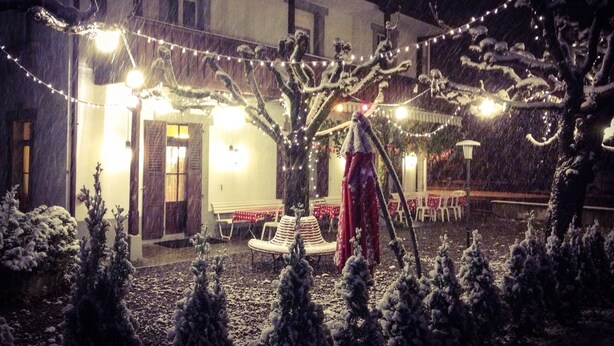 Un hotel llamado 'HOTEL ALPINA' con un jardín cubierto de nieve, luces de adorno y mobiliario exterior.