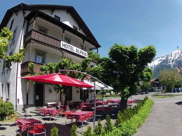 Un hotel llamado "HOTEL ALPINA" con mesas al aire libre y montañas al fondo.