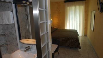 Baño con lavamanos, espejo y ducha. Se observa una cama con colcha estampada y una ventana con cortinas.