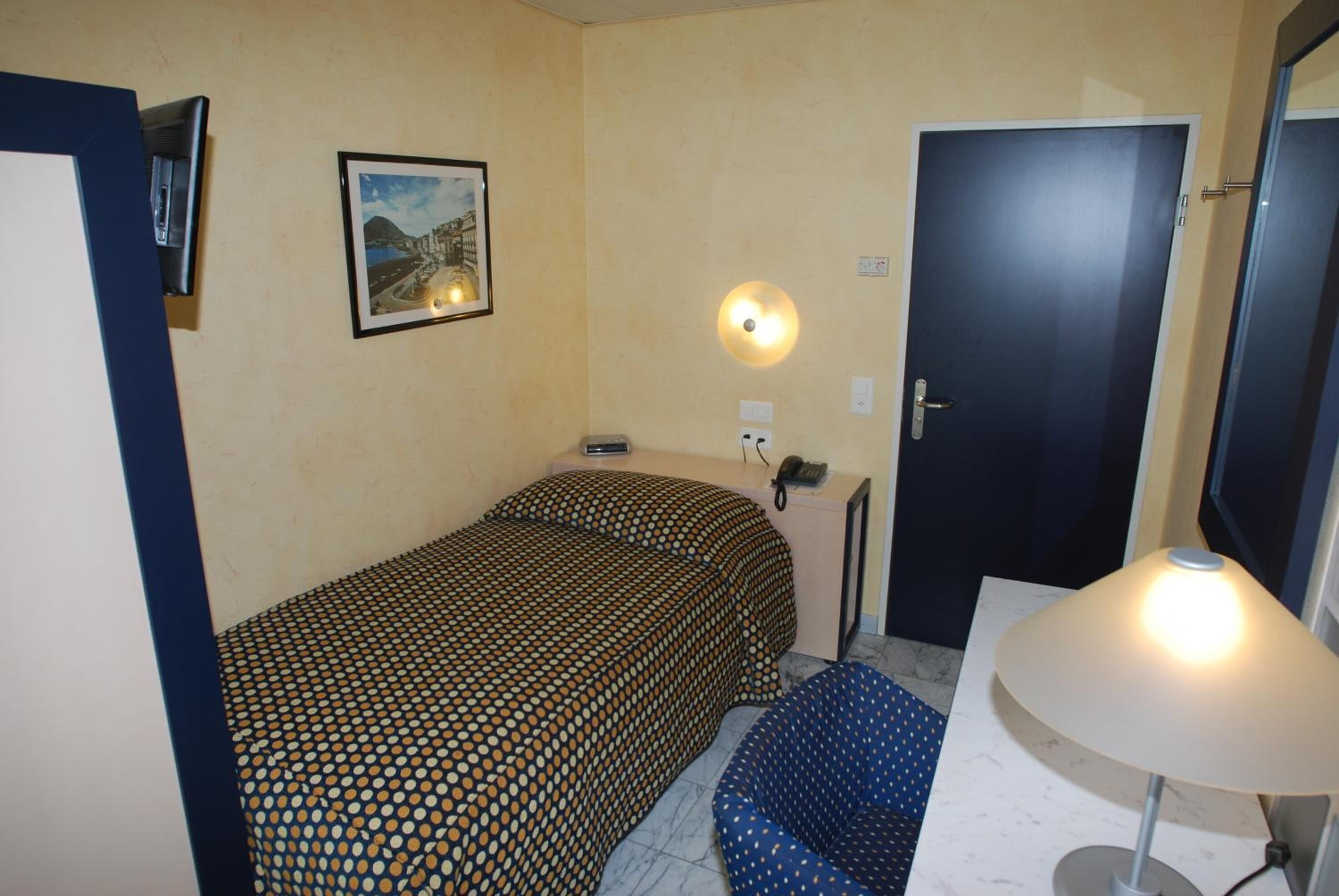 Ein kleiner, gemütlicher Raum mit einem Bett, einem Sessel, einem Schreibtisch mit Lampe, einem an der Wand montierten Telefon und einer Tür.