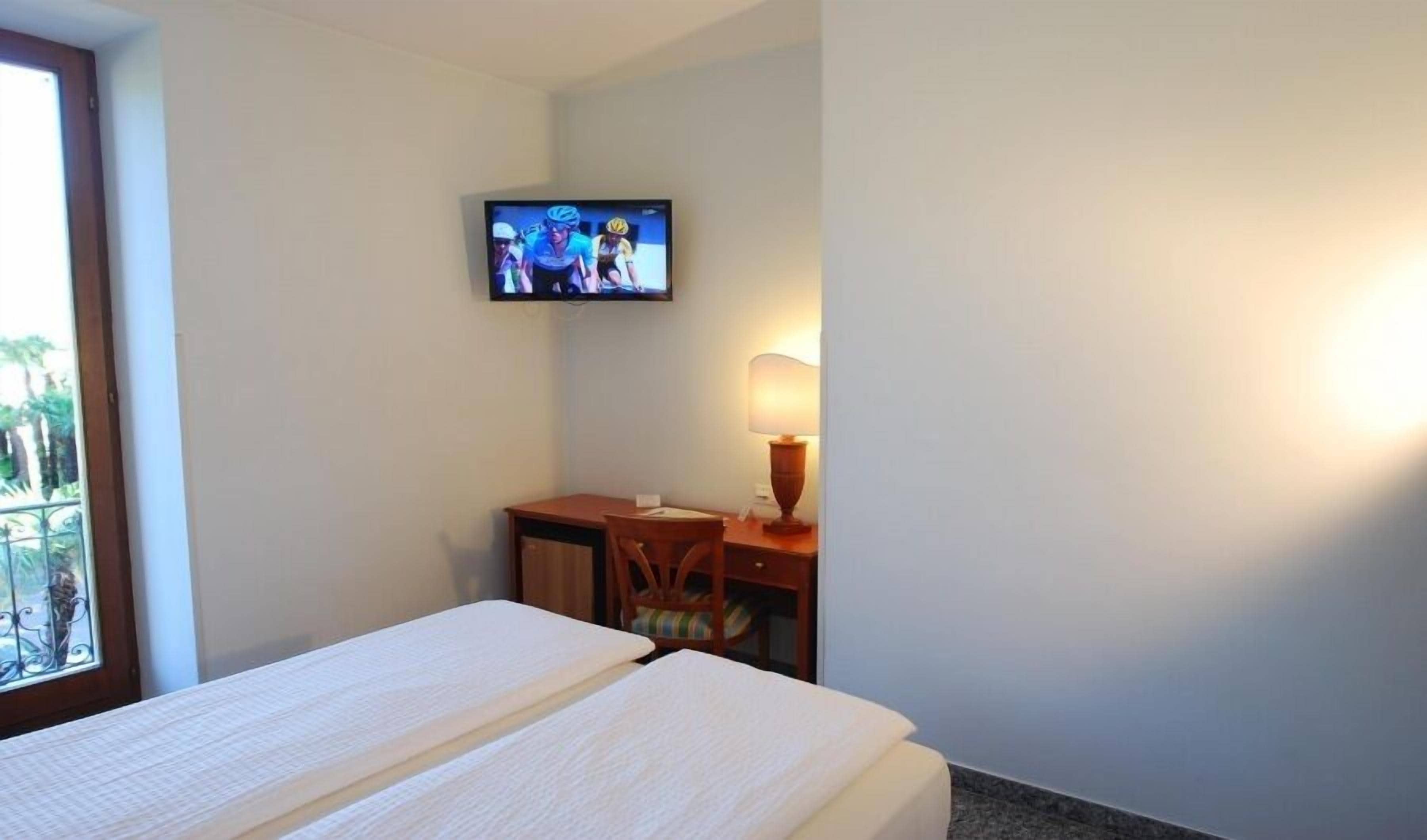A hotel room with two beds, a desk, a chair, a TV on the wall, and a window with a view of palm trees.