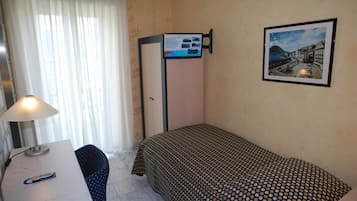 Una habitación de hotel pequeña con una cama individual, un escritorio con lámpara, una silla, un cuadro en la pared y un cartel con información.