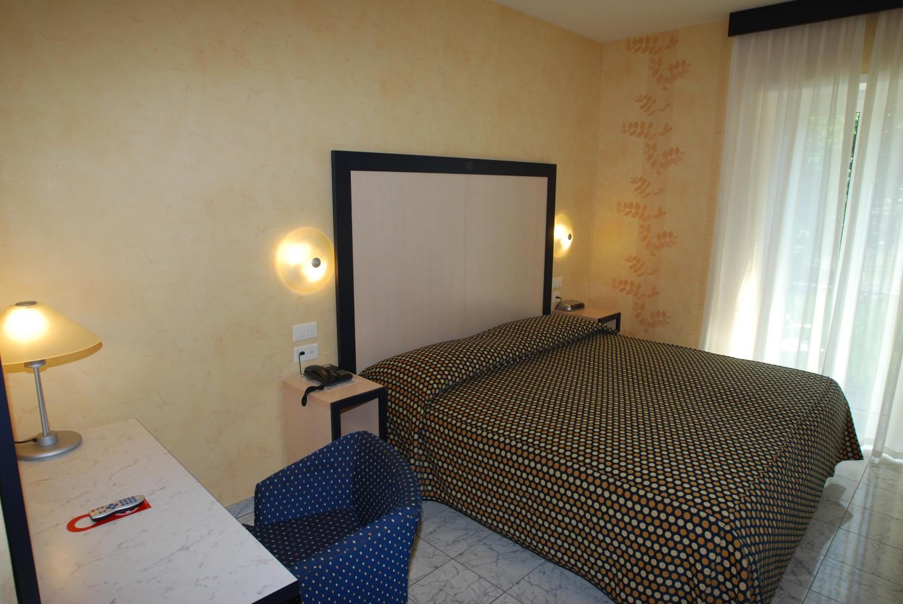 Una camera d'albergo con un letto, una scrivania con una lampada, una sedia e una finestra con tende.